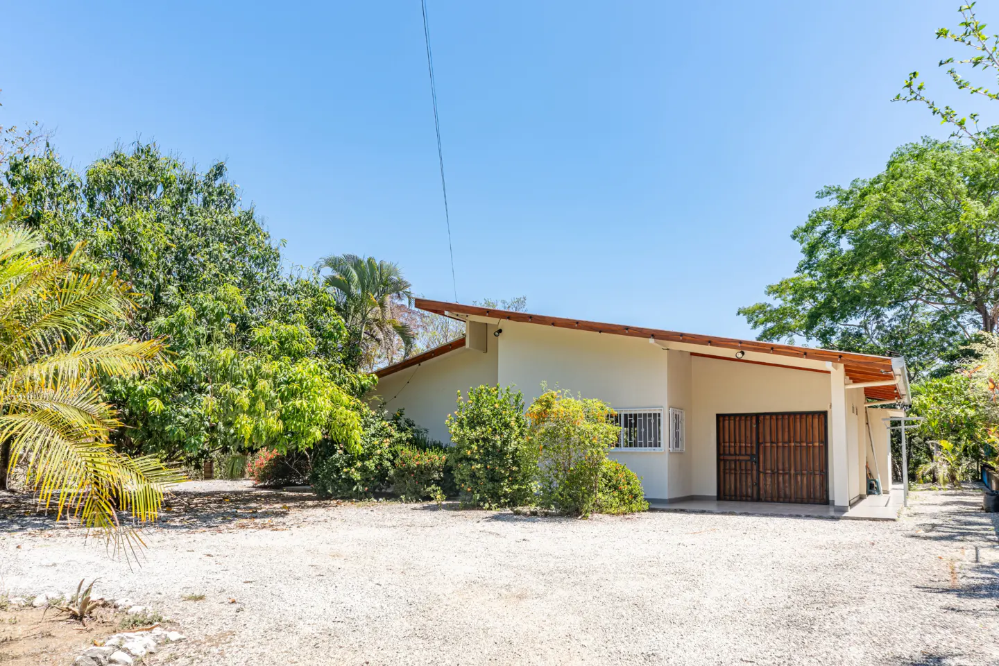 Beige single-story house with a brown roof and a double garage door surrounded by green trees under a clear blue sky.