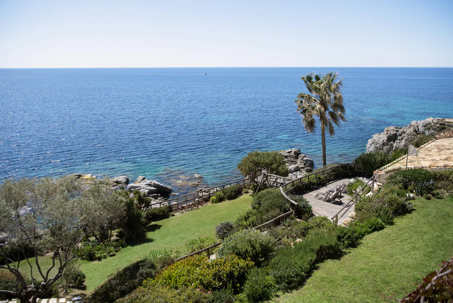 Ocean view property with a palm tree, green lawn, and wooden deck with lounge chairs. Blue sea and sky.
