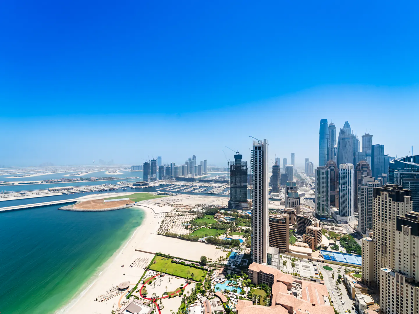 Aerial view of the Dubai coastline with modern skyscrapers, a sandy beach, and turquoise water under a clear blue sky.