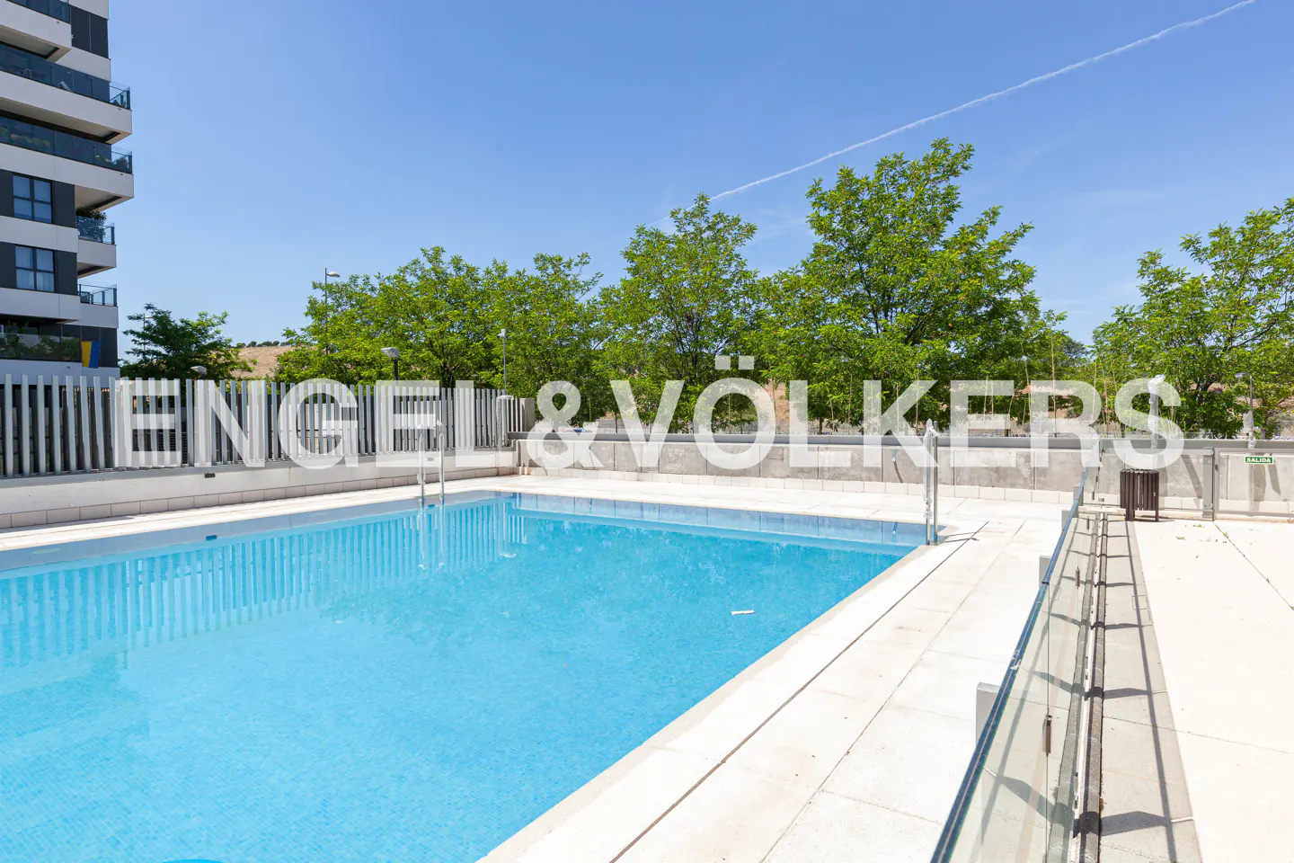 Outdoor pool with blue water, surrounded by a white tiled deck and green trees under a blue sky. Engel & Völkers logo overlay.