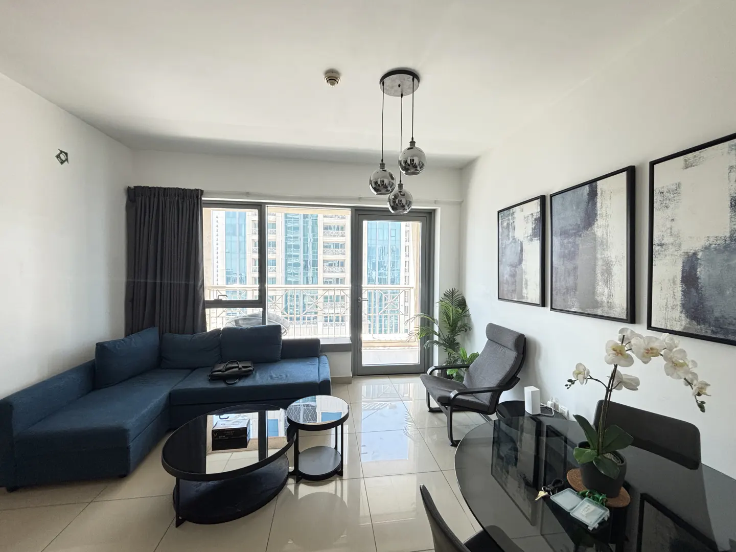 Bright living room with blue sofa, black tables, and city view through large windows. Modern art and a dining area complete the space.