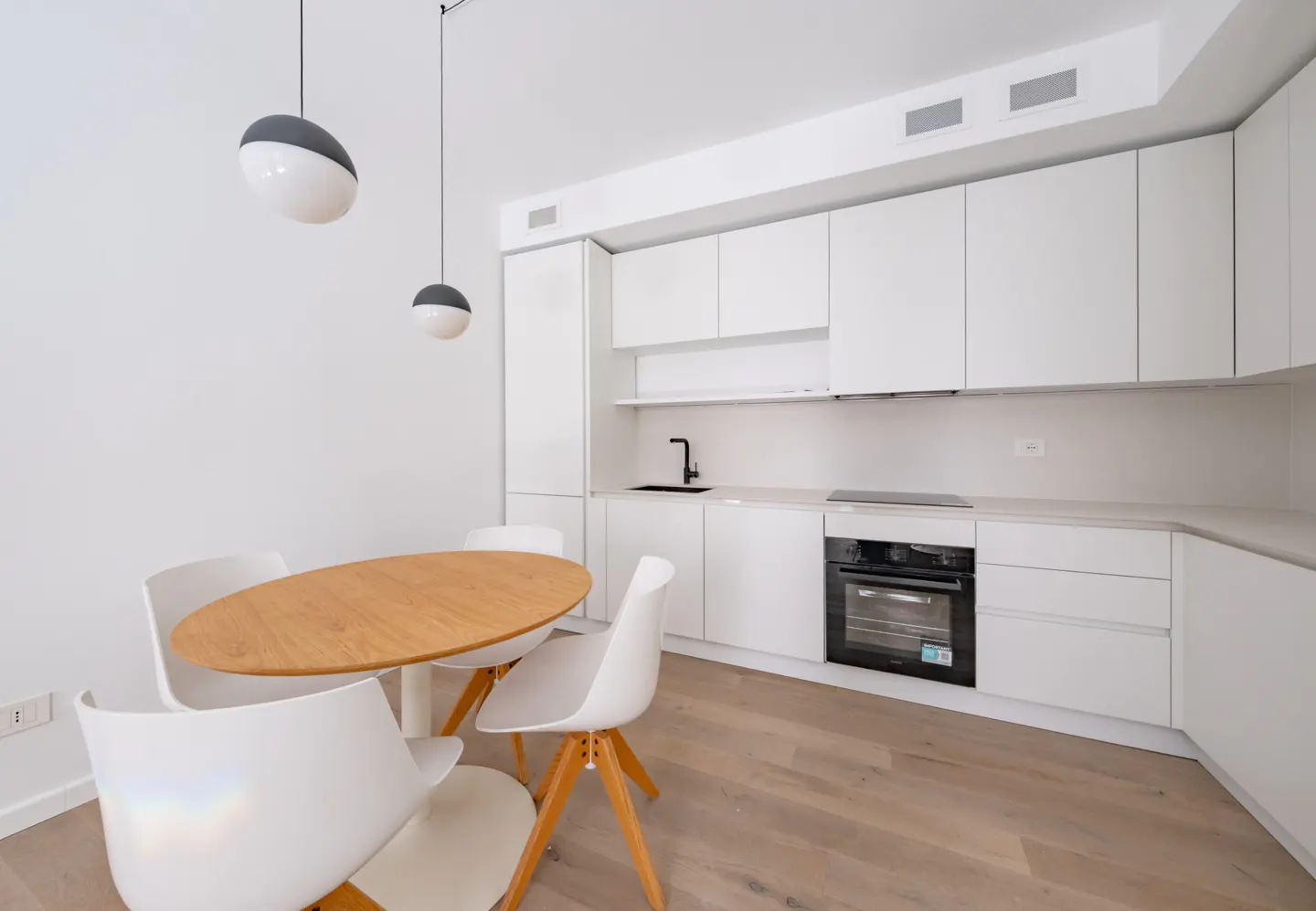 Bright, modern kitchen with white cabinets, wood floors, and a round wood table with white chairs. Two pendant lights hang above.