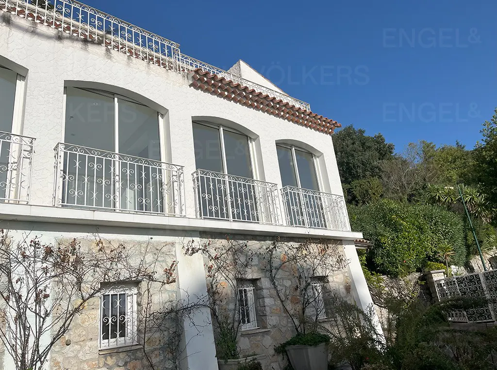 Exterior view of a white stucco house with arched windows, white railings, and a stone foundation with climbing vines. Blue sky background.