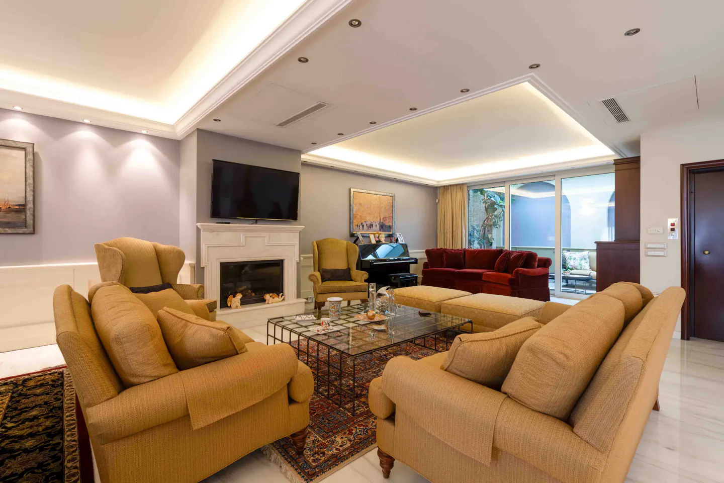 Living room with tan sofas, fireplace, TV, piano, and red sofa. A glass coffee table sits on a patterned rug.
