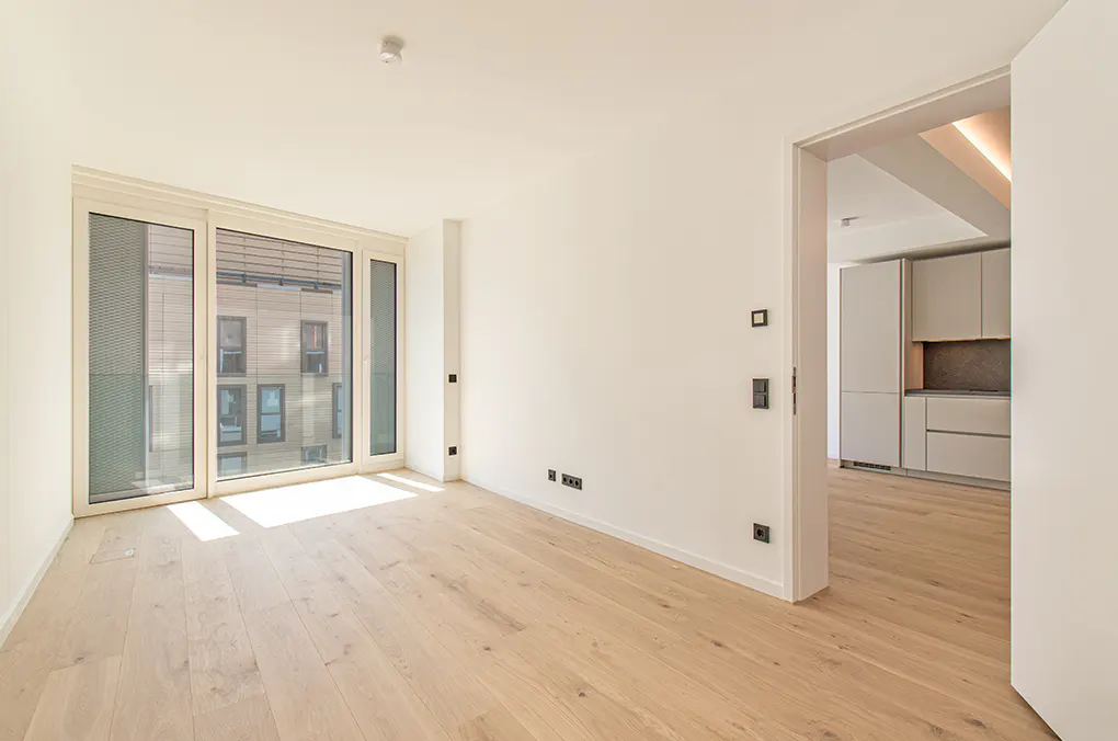 Bright, empty room with light wood floors, large windows, and a doorway leading to a modern kitchen with grey cabinets.