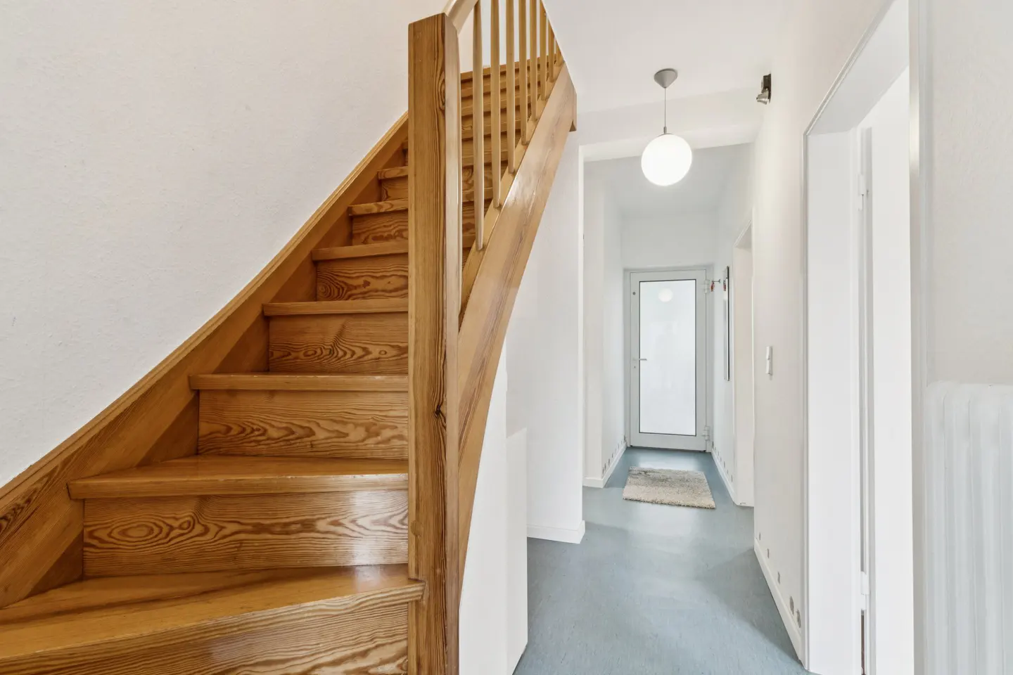 Interior view of a home featuring a wooden staircase, white walls, and a hallway with a door at the end.