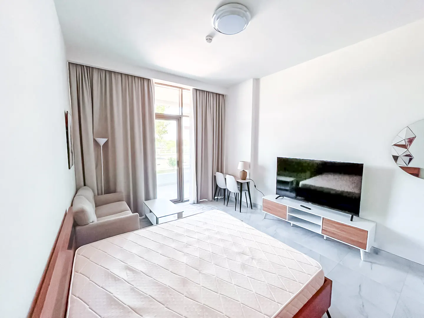 Modern hotel room with a bed, small sofa, TV on a wooden stand, desk with two chairs, and floor-to-ceiling beige curtains.