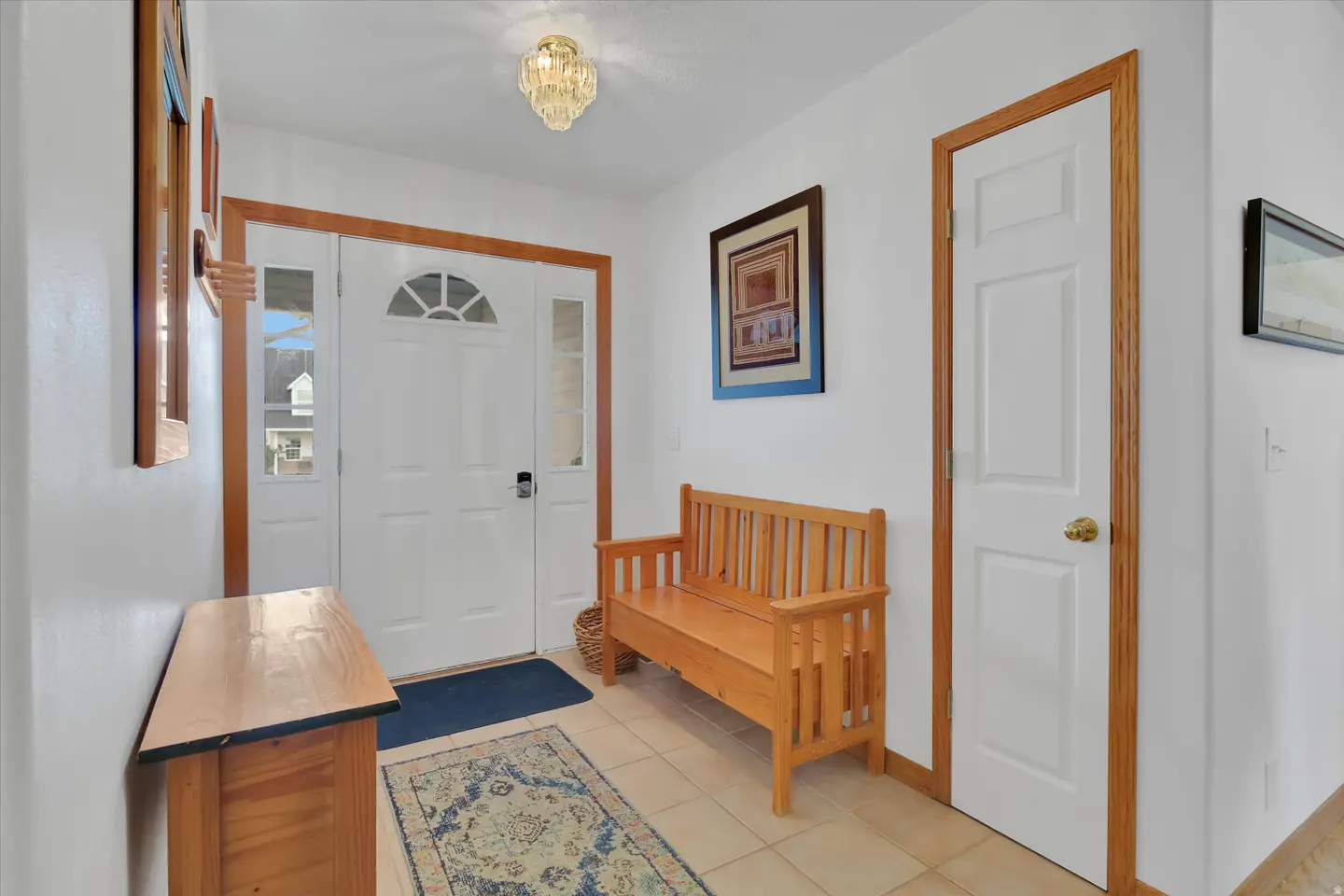 Bright foyer with white walls, tan tile floor, and wood trim. A wooden bench sits near the front door, with a blue rug on the floor.