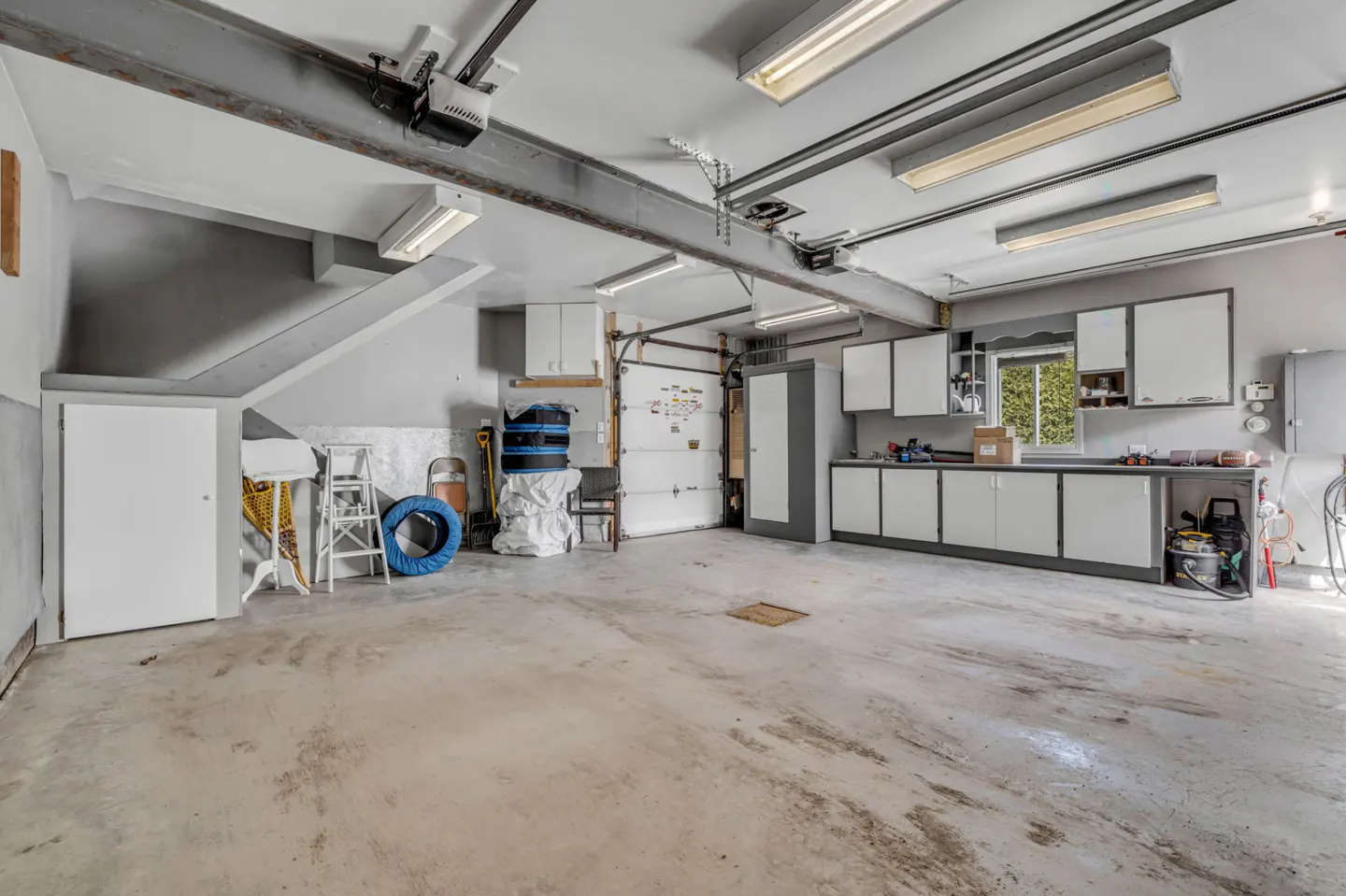 A well-lit garage with gray walls, a concrete floor, and white cabinets. A garage door is partially open. Tools and storage items are visible.
