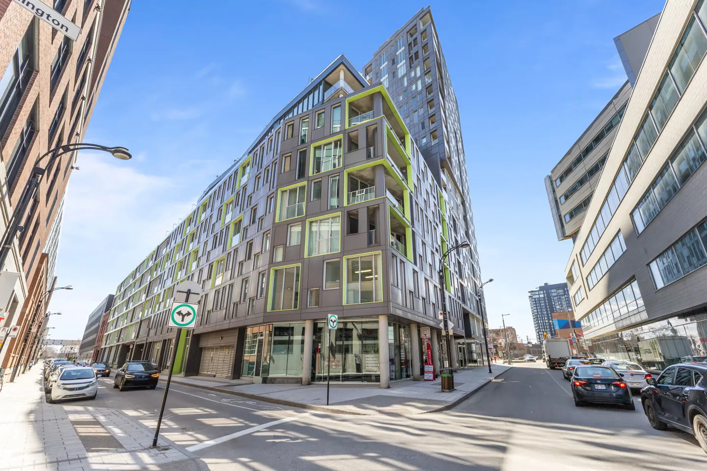 Street view of a modern gray building with green window frames under a blue sky. Cars are parked along the street.