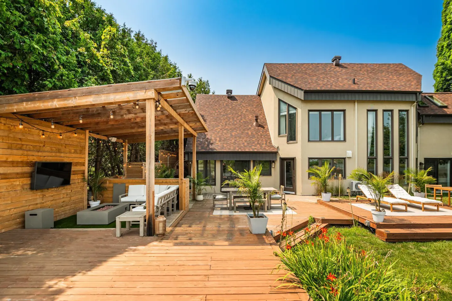 A backyard with a wooden deck, a pergola with string lights, and a modern house with large windows.