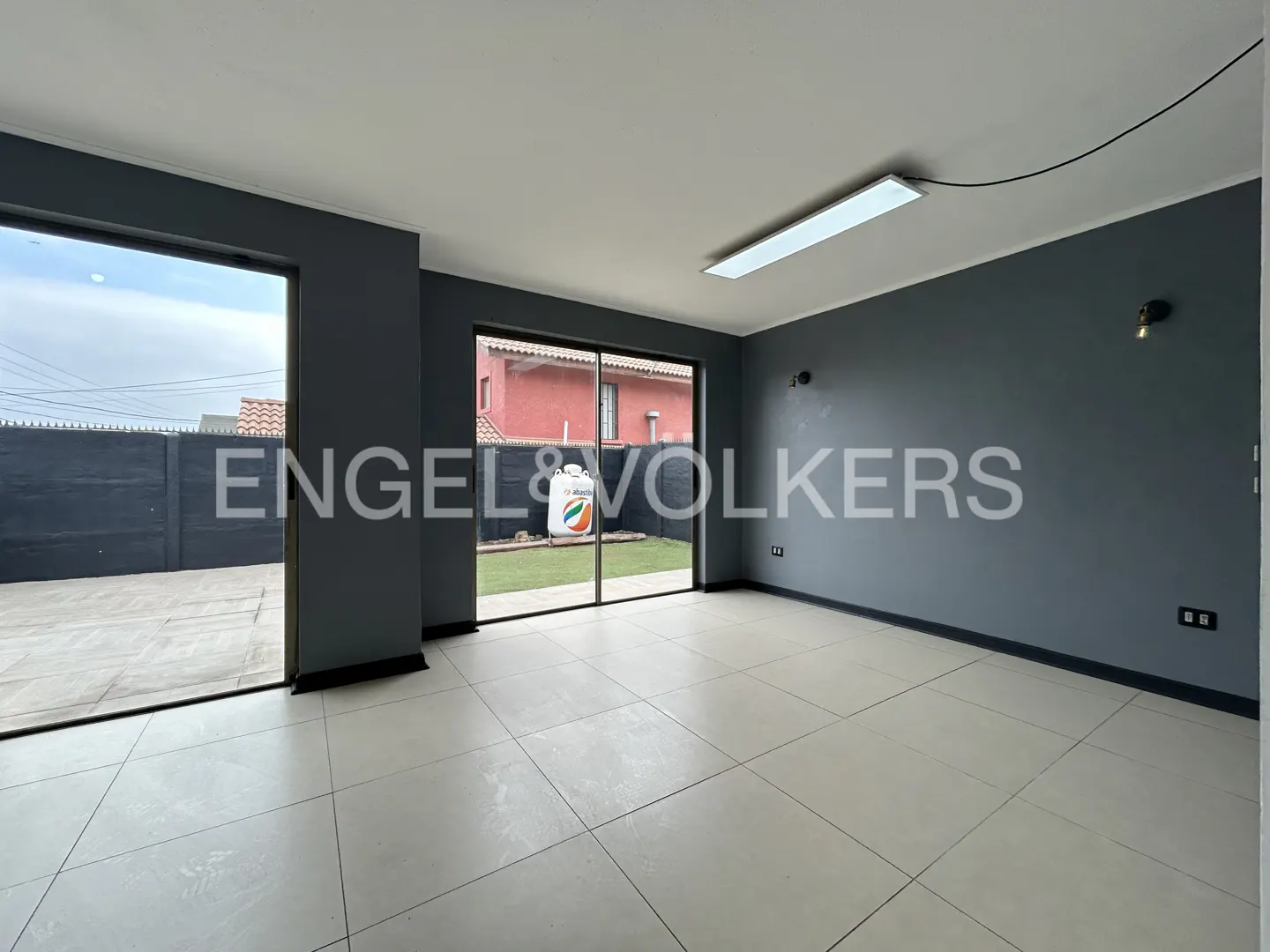 Empty room with grey walls, large sliding glass doors, light tile floors, and a view of a patio and red house exterior.