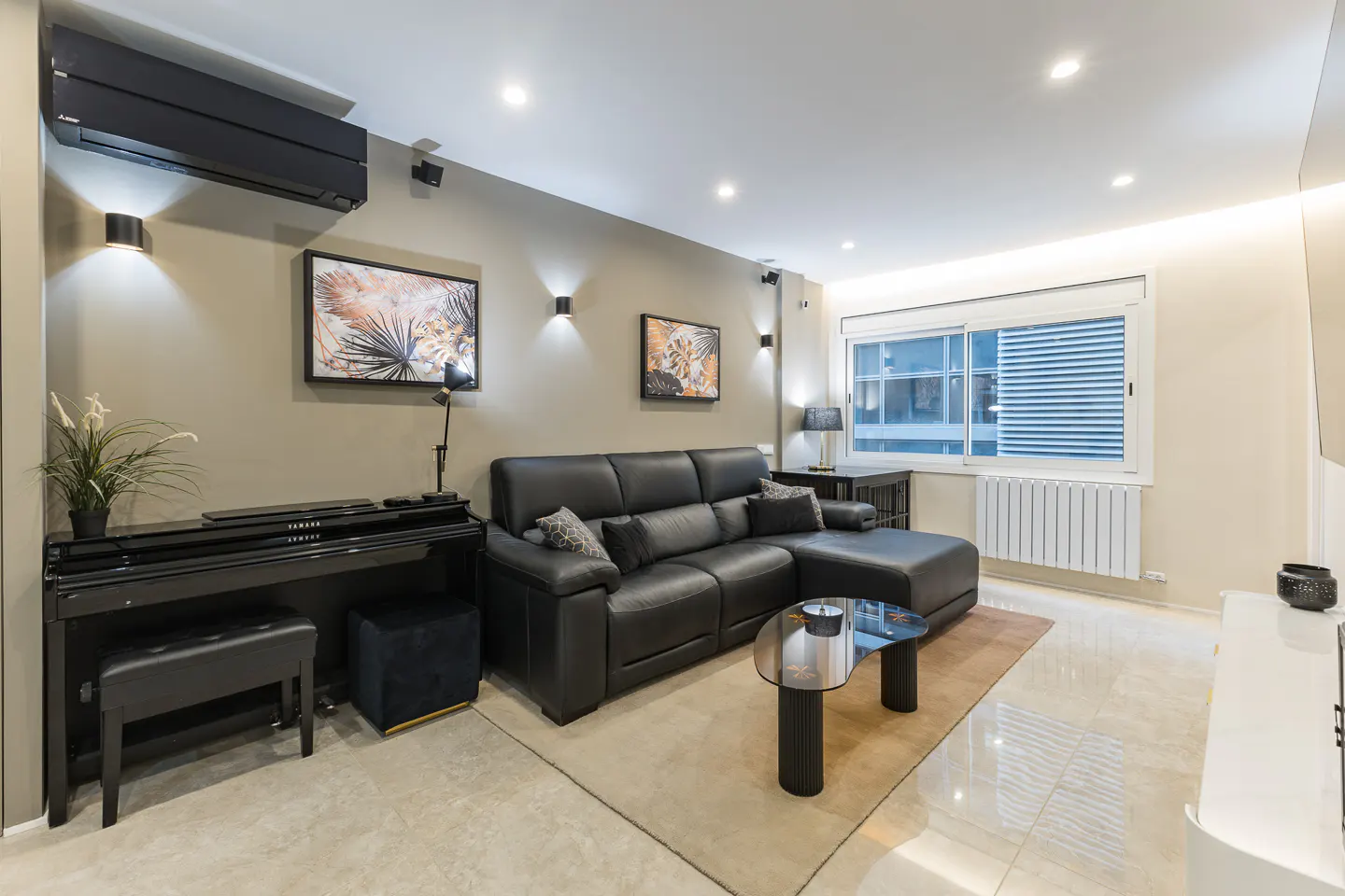 A modern living room with a black leather sofa, piano, and glass coffee table on a beige rug. Two framed art pieces hang on the wall.
