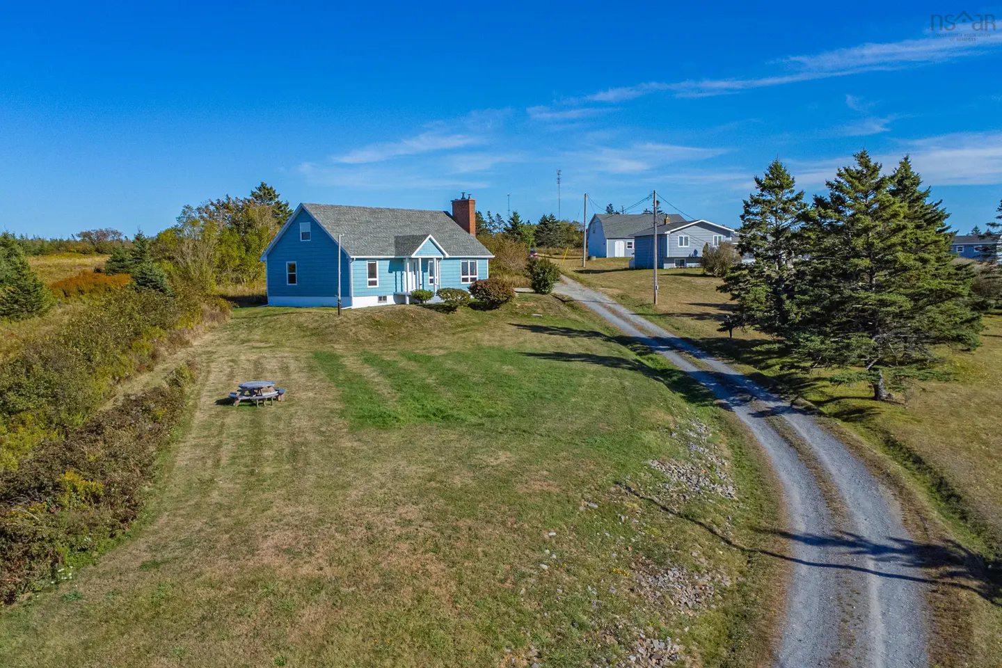 Aerial view of a light blue house with a gray roof and brick chimney, set on a grassy lawn under a clear blue sky. A gravel driveway leads to another house in the distance.