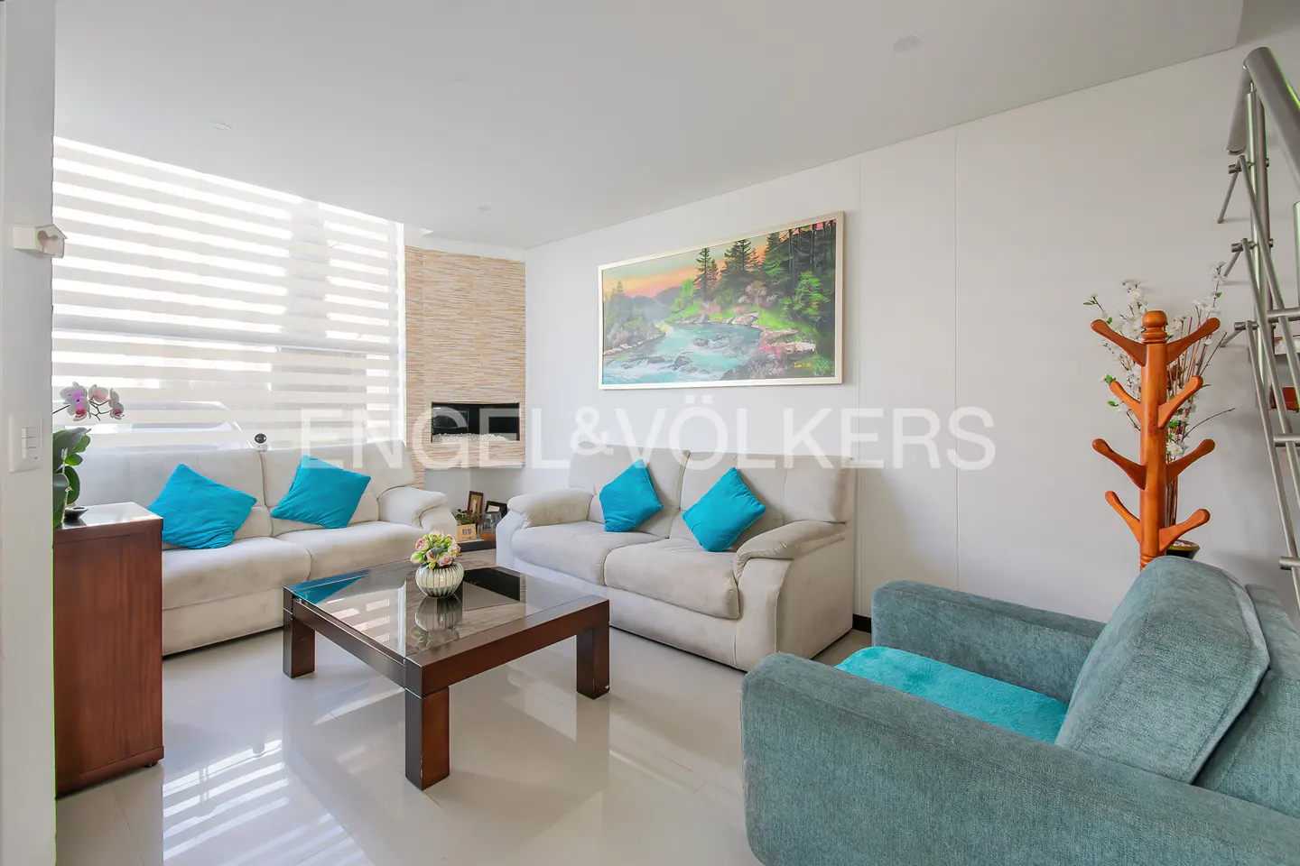 Bright living room with two sofas, blue pillows, a glass coffee table, and a blue armchair. A painting hangs above the sofa.