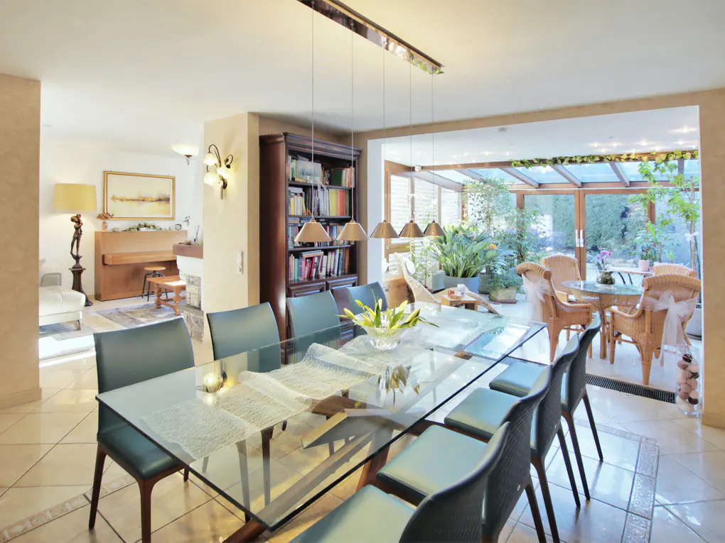 Modern dining room with a glass table, dark chairs, and a view into a sunlit conservatory filled with plants.