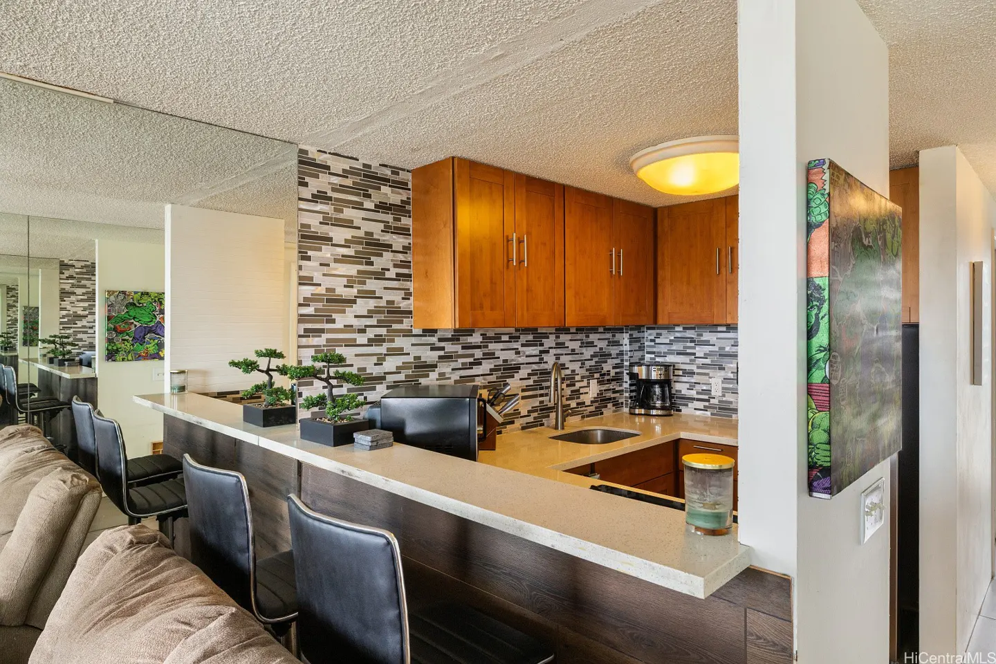 A kitchen with wood cabinets, mosaic tile backsplash, and a white countertop bar with black stools. A bonsai tree sits on the counter.