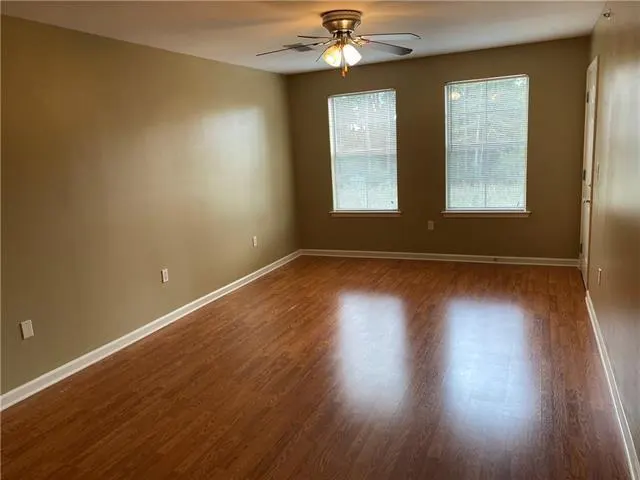 Empty room with tan walls, wood-look flooring, two windows with blinds, and a ceiling fan.