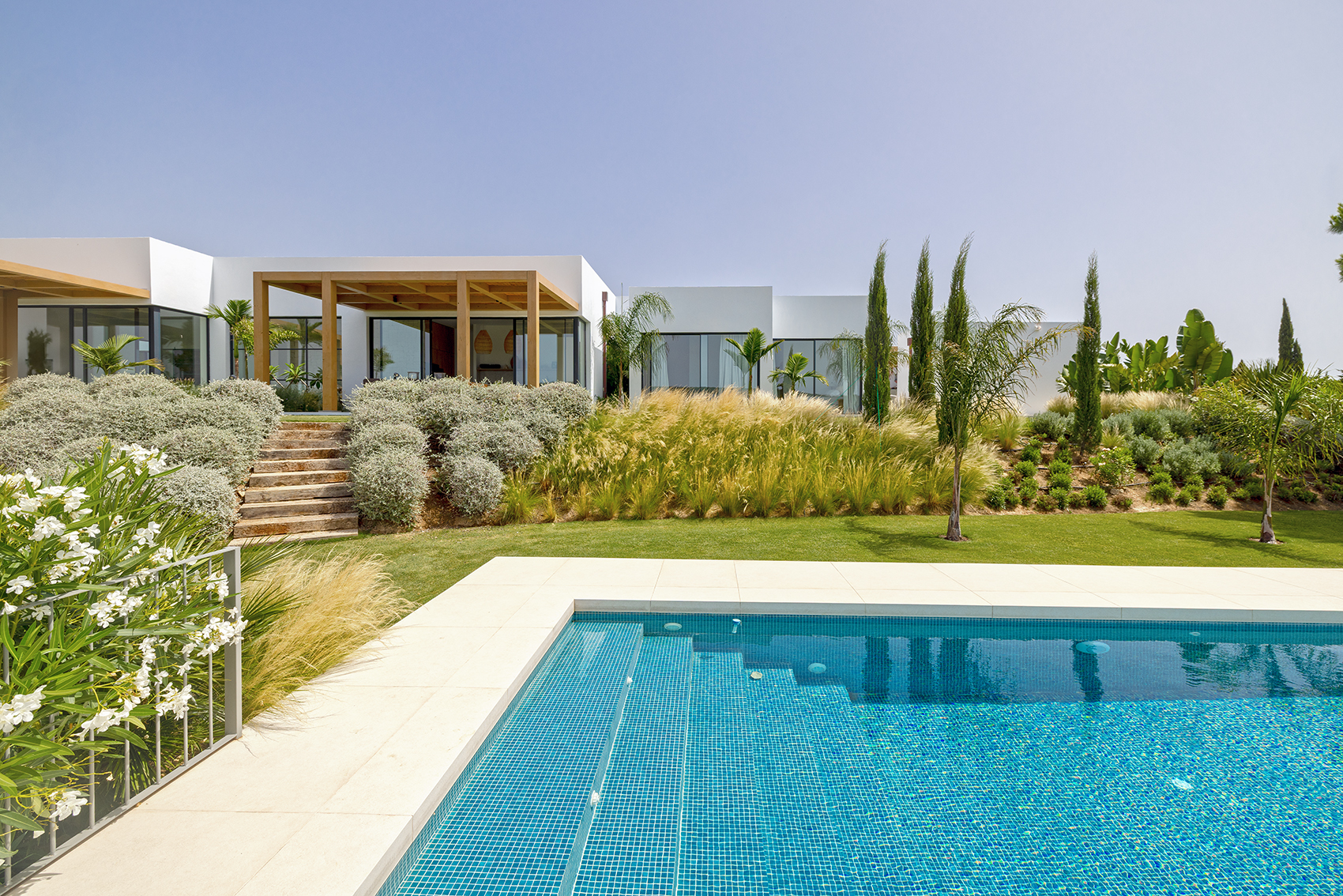 Modern white villa with a blue tiled pool, stone steps, and lush landscaping under a clear blue sky.