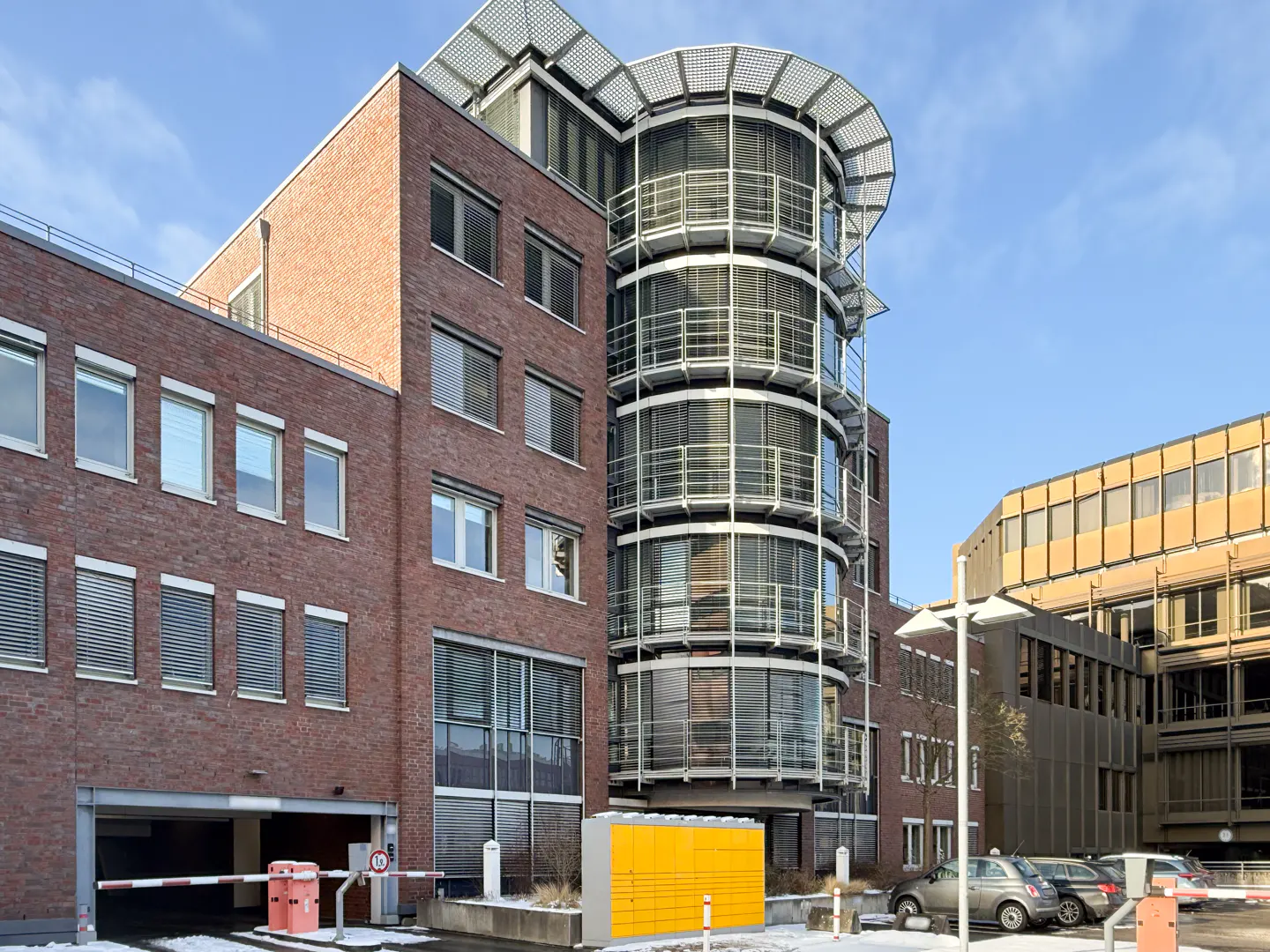 Exterior view of a brick office building with a cylindrical glass tower and a parking garage entrance.
