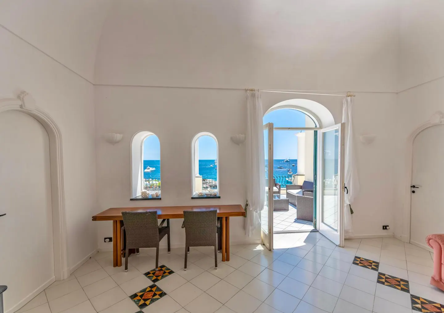 Bright room with arched windows and doors. A wooden table and chairs face the ocean view. Tiled floor and white walls.