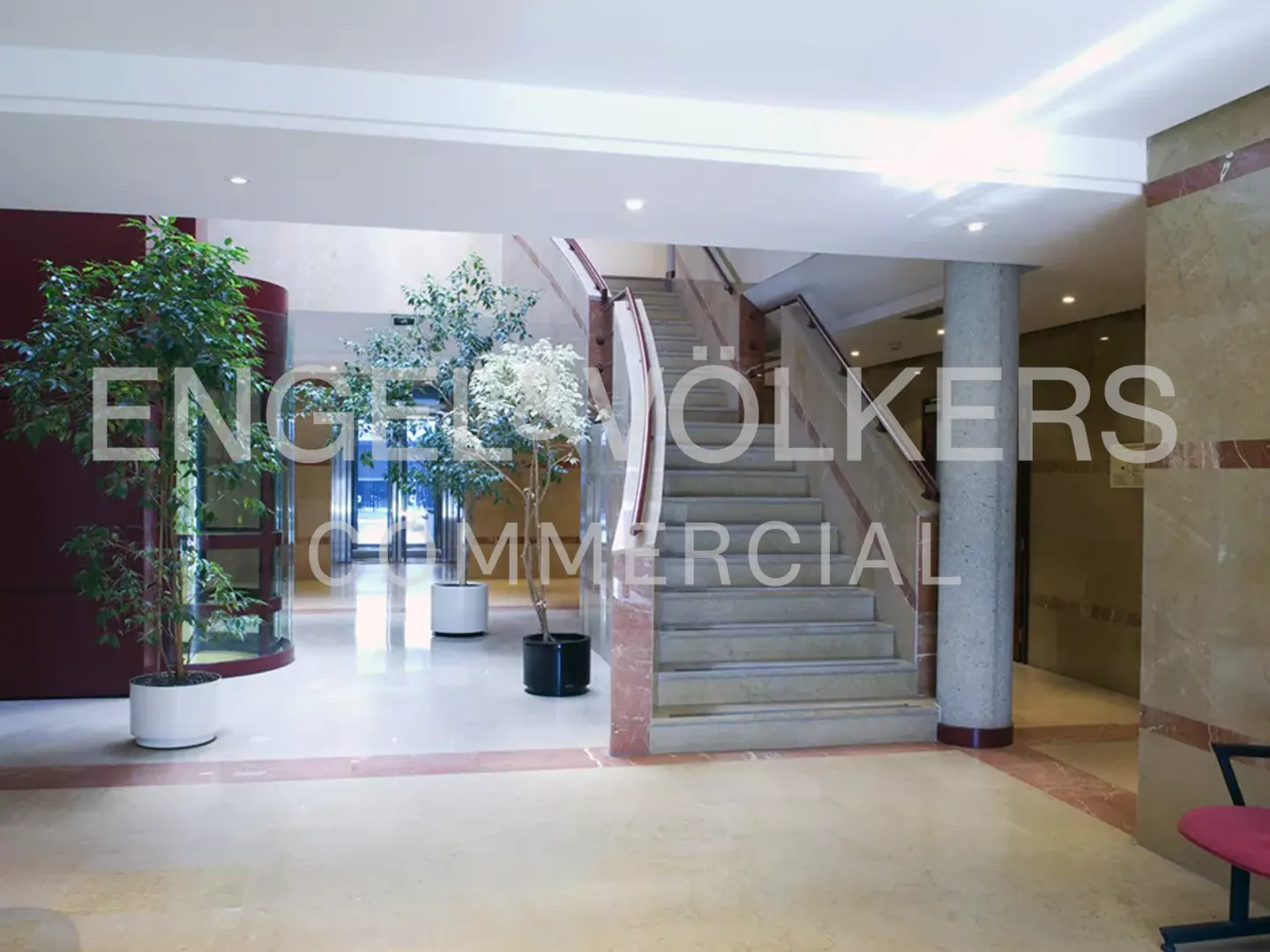 Bright lobby with marble floors, potted trees, and a staircase with marble accents. Natural light filters through glass doors.