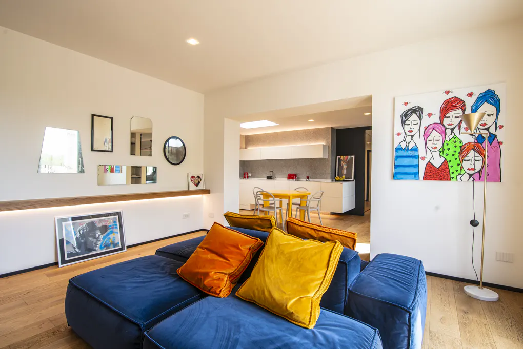 Bright living room with blue sofa, orange and yellow pillows, art, mirrors, and a view into a modern kitchen with a yellow table.