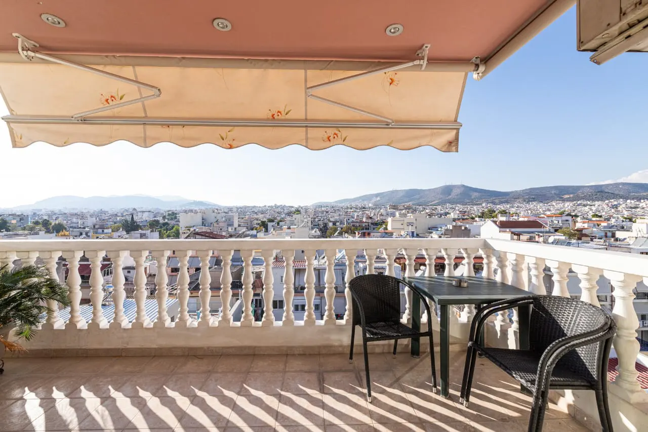 Balcony with white balustrade, table, and chairs overlooking a city with mountains in the background under a beige awning.
