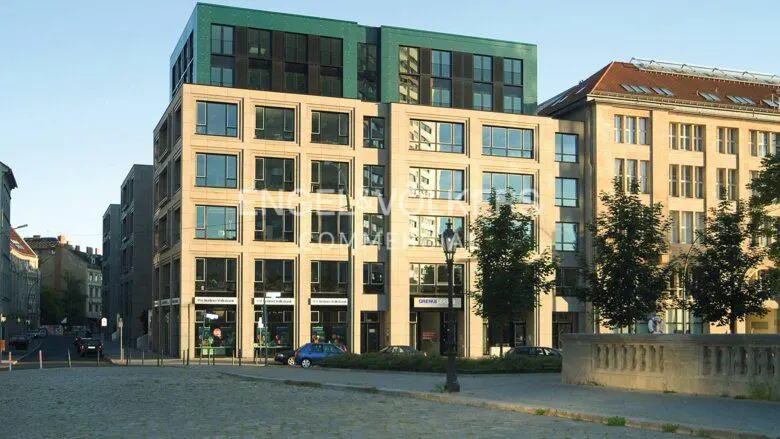 A modern, multi-story building with a green rooftop and large windows, situated on a cobblestone street.