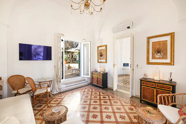Bright living room with white walls, patterned tile floor, and an open door to a patio. Wicker furniture and a chandelier add charm.