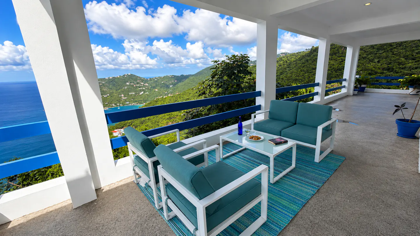 Outdoor patio with turquoise furniture, white frames, and blue railings overlooking a lush green hillside and ocean view.
