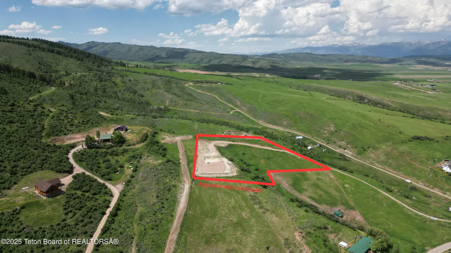 Aerial view of a green, hilly landscape with a red outline marking a lot. Mountains are visible in the background under a cloudy sky.