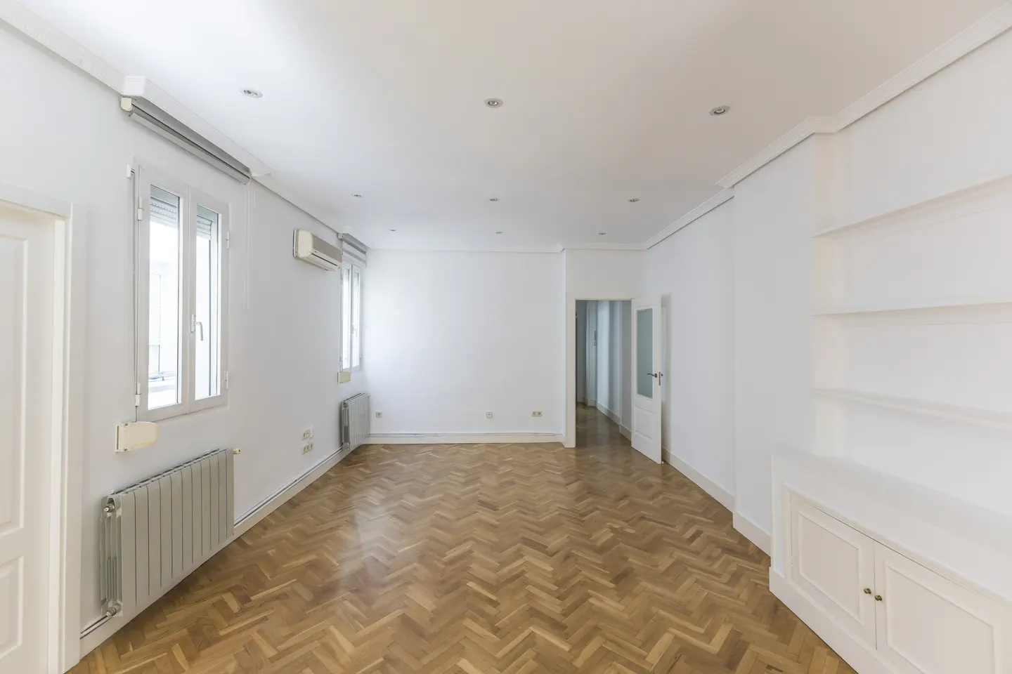 Bright, empty room with herringbone wood floors, white walls, windows, and built-in shelves.
