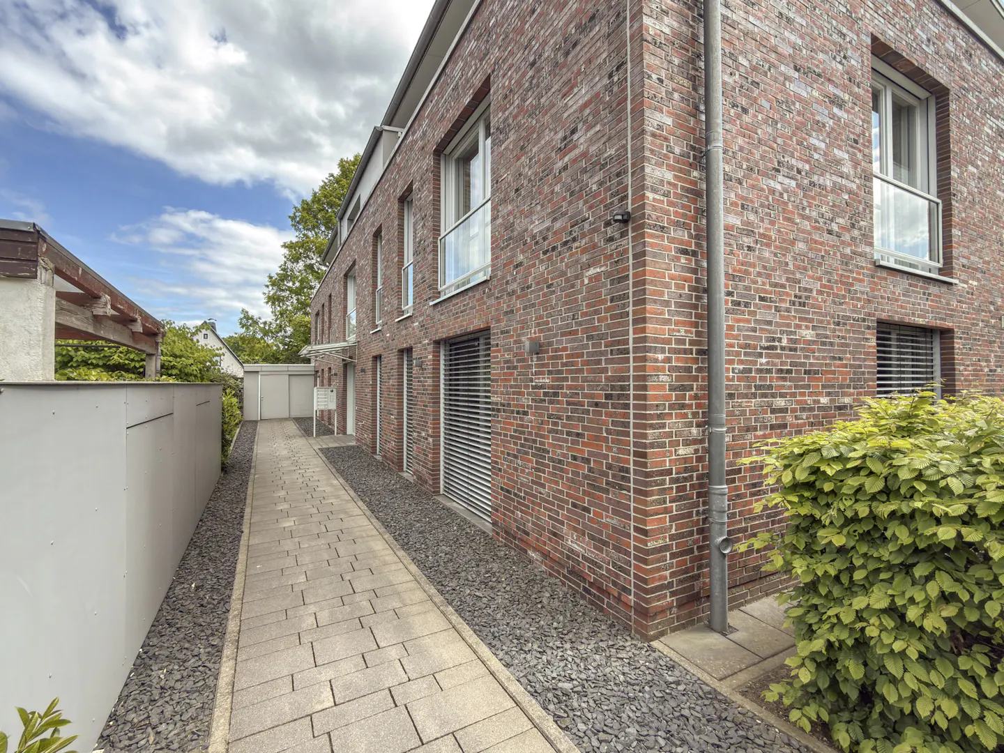 A paved walkway leads along the side of a modern red brick apartment building with large windows and a green bush.