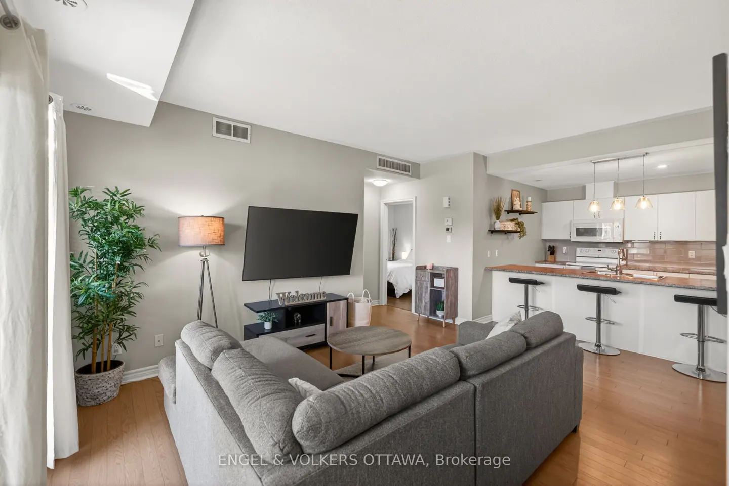Open-concept living space with a gray sectional sofa, TV, and kitchen island with bar stools. Hardwood floors and neutral wall colors.