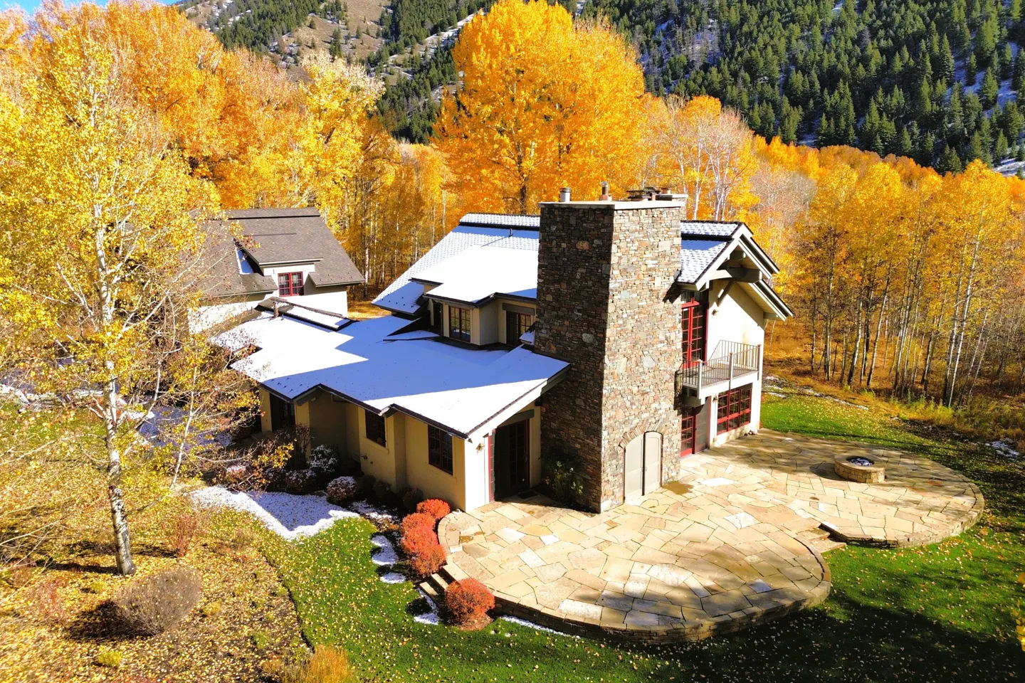 Aerial view of a tan house with a stone chimney, snow on the roof, and fall foliage in the background.