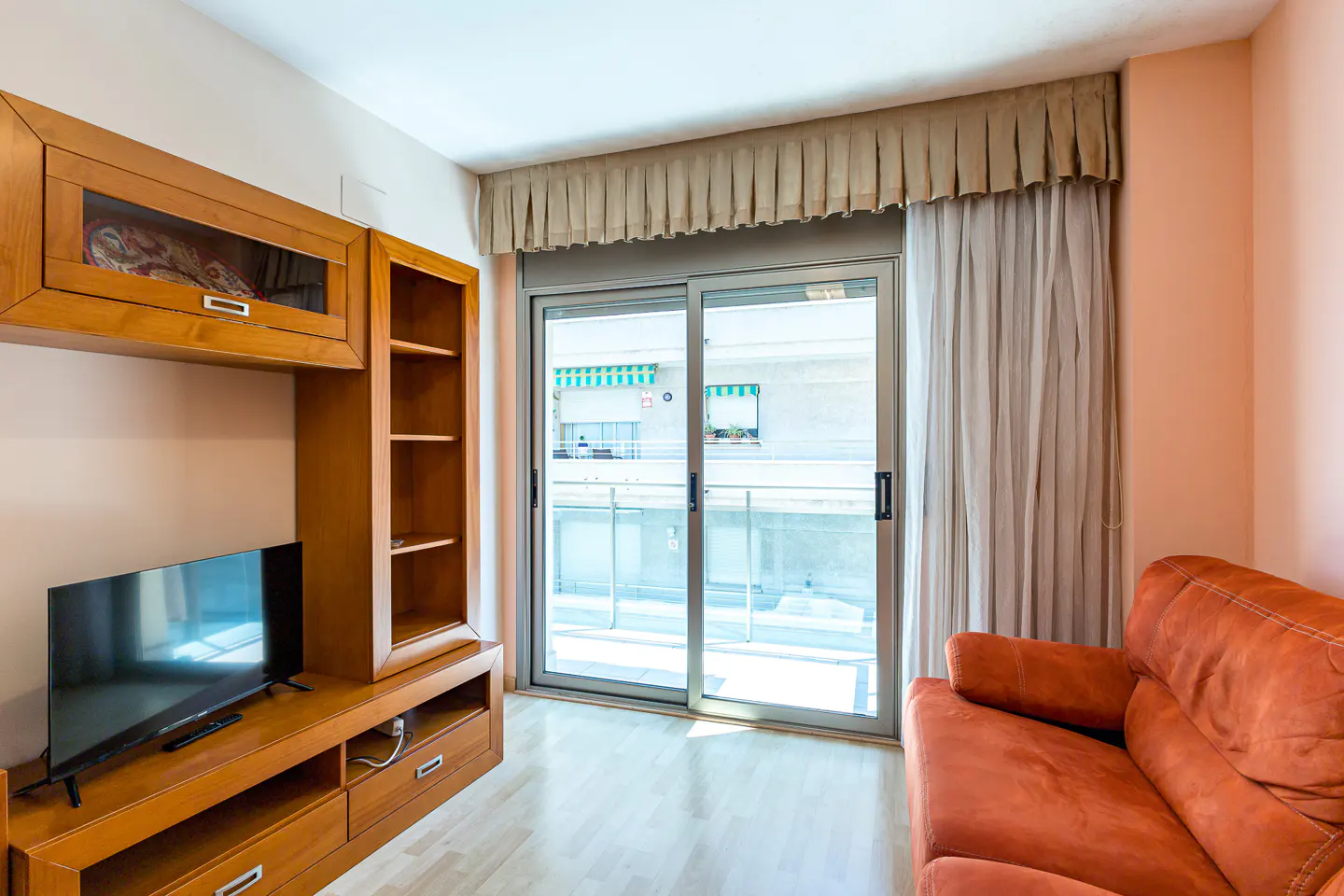 Living room with a wooden entertainment unit, flat-screen TV, orange sofa, and sliding glass door leading to a balcony.