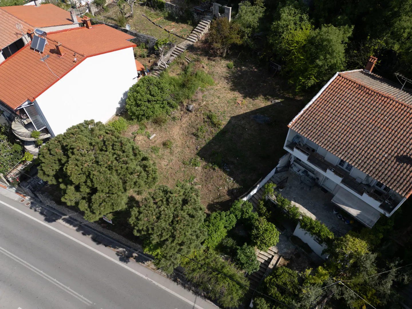 Aerial view of a vacant lot between two houses with red tile roofs and green trees.