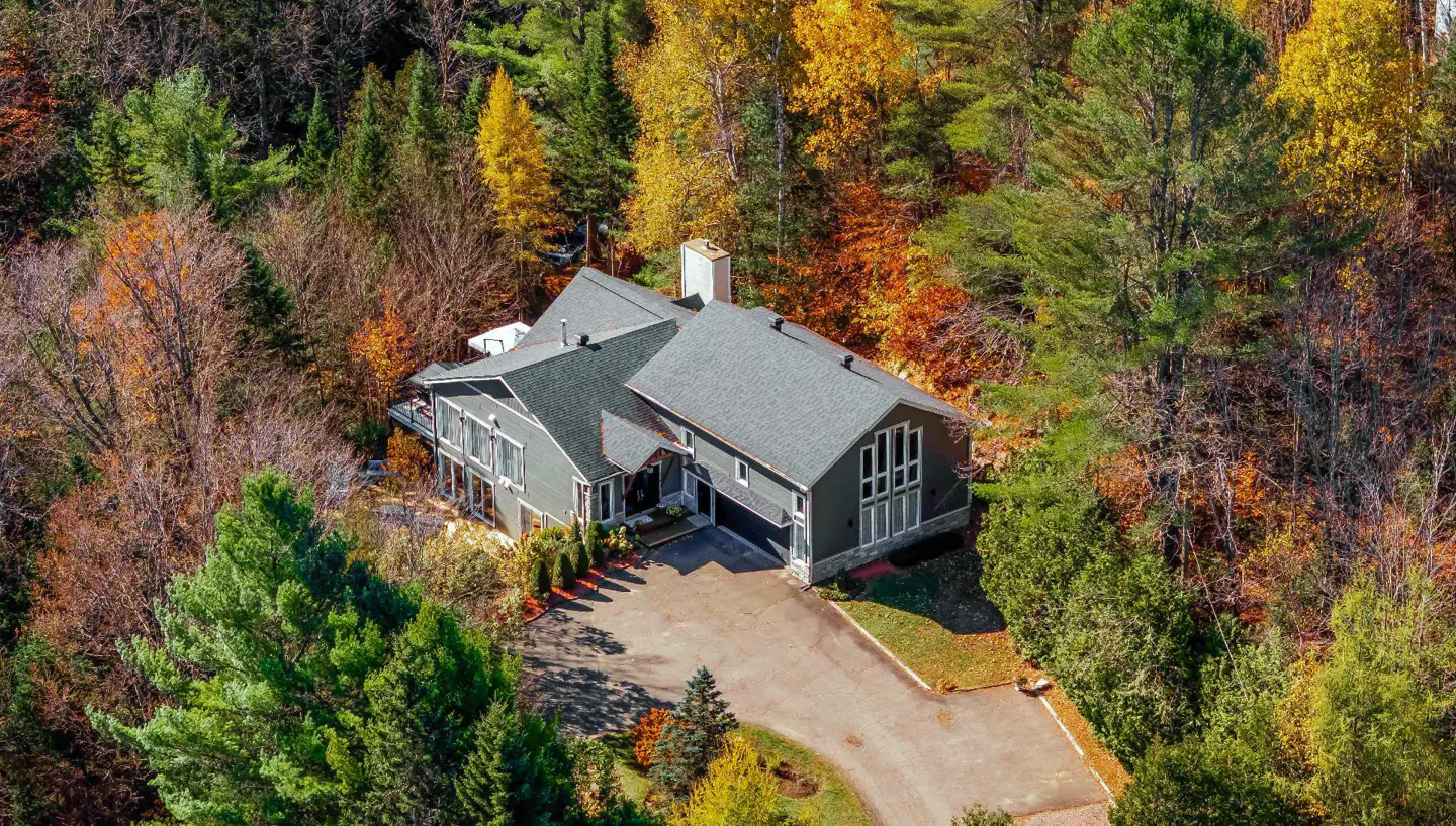 Aerial view of a modern gray house with a gray roof, surrounded by colorful autumn trees. A driveway leads to the house.