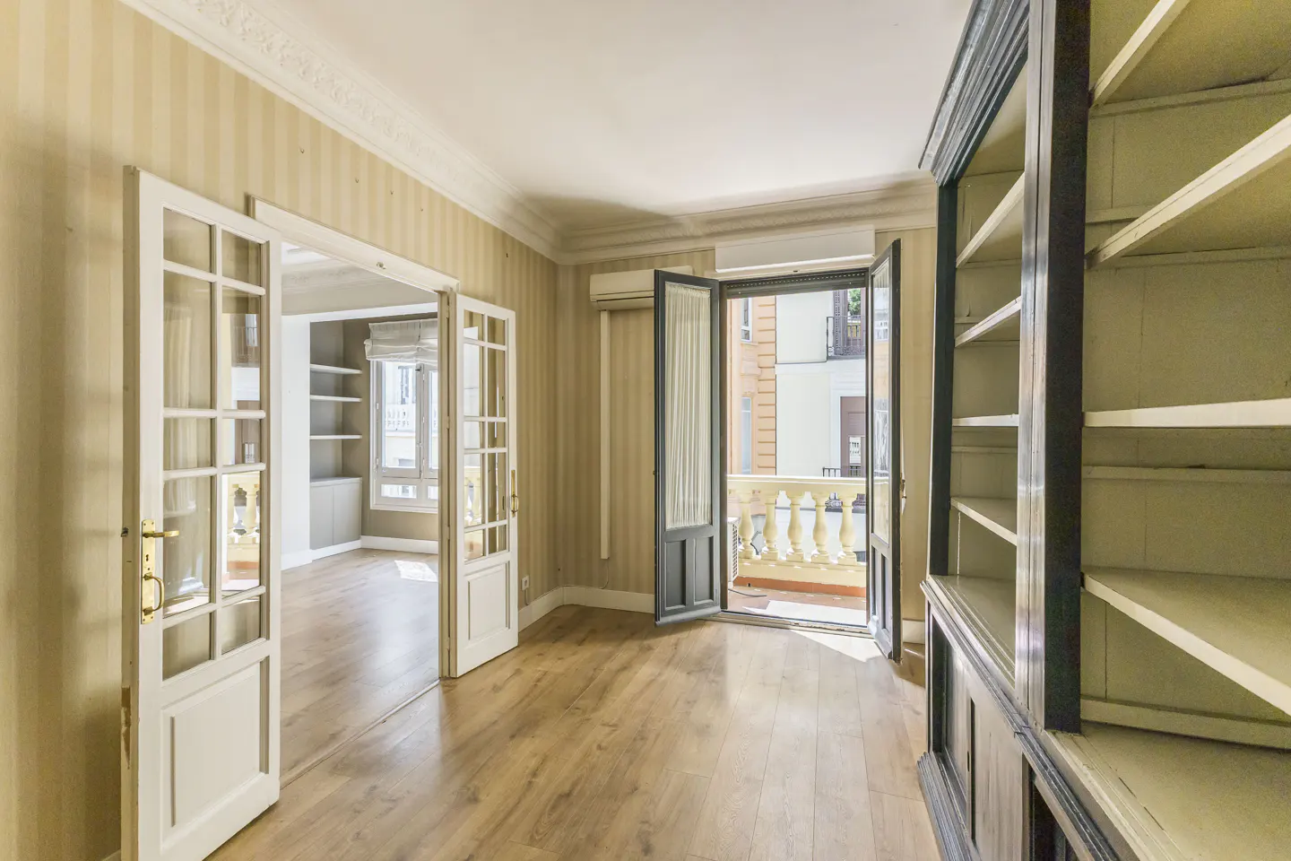 A bright room with wood floors, striped wallpaper, and white trim. Open glass doors lead to another room and a balcony. A large, dark bookcase stands on the right.