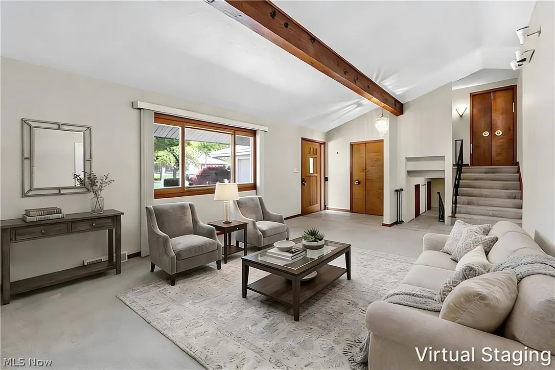 A bright living room with a beige sofa, two gray chairs, and a wooden coffee table on a patterned rug. Stairs lead to double doors.