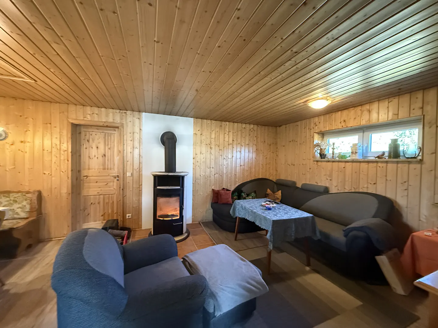 Cozy wood-paneled living room with a black wood-burning stove, blue armchair, and gray sectional sofa. A window provides natural light.