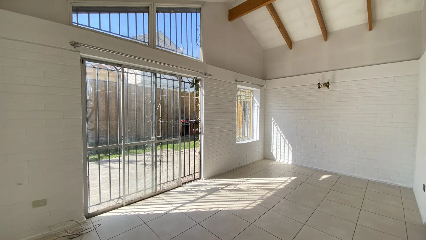 Bright, empty room with white brick walls, tile floor, and vaulted ceiling with exposed beams. Metal bars cover the windows and sliding glass door.