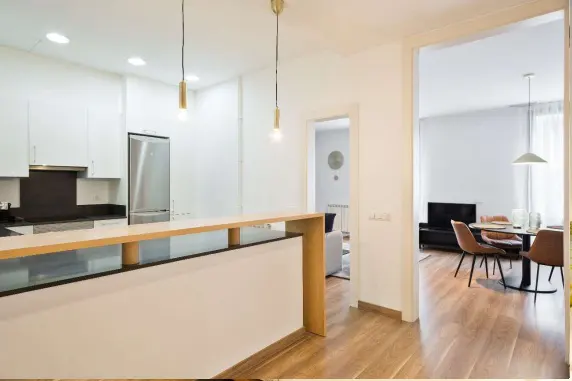 Bright, modern kitchen with white cabinets, stainless steel refrigerator, and wood floors. A doorway leads to a dining area with a table and chairs.