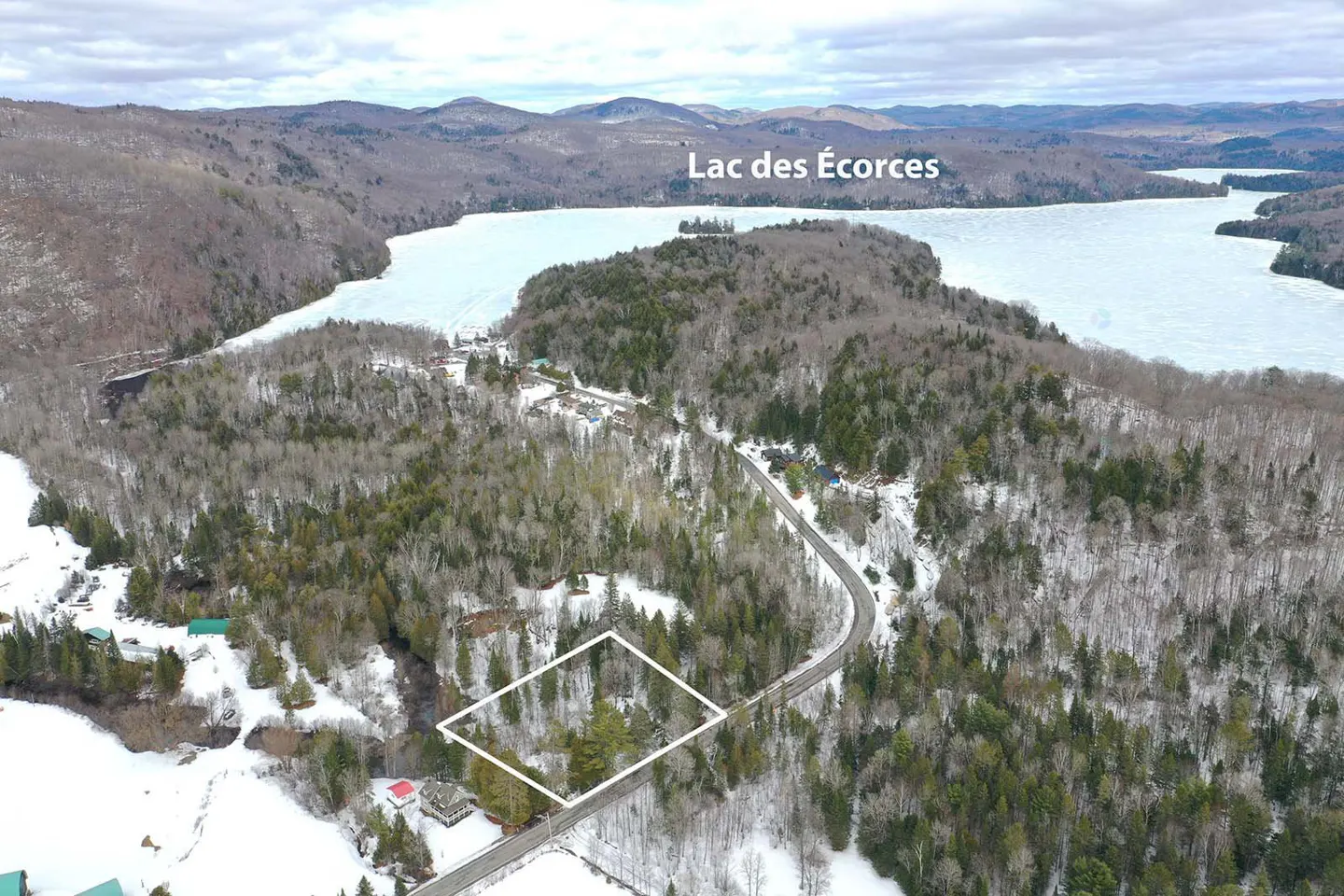 Aerial view of a wooded lot outlined in white, near Lac des Écorces, with snow-covered ground and mountains in the background.