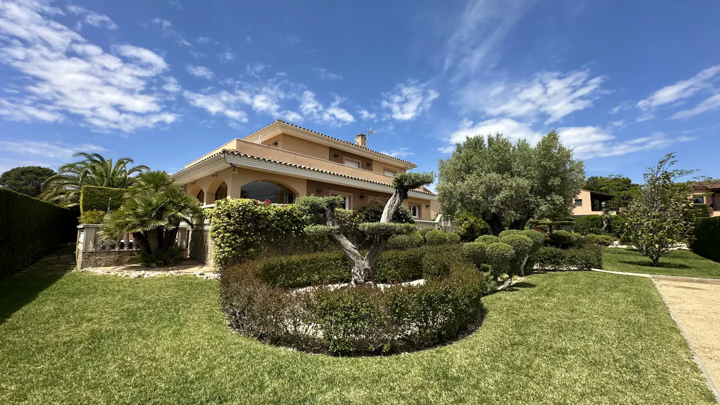 Two-story tan house with a manicured lawn and garden under a blue, cloudy sky.