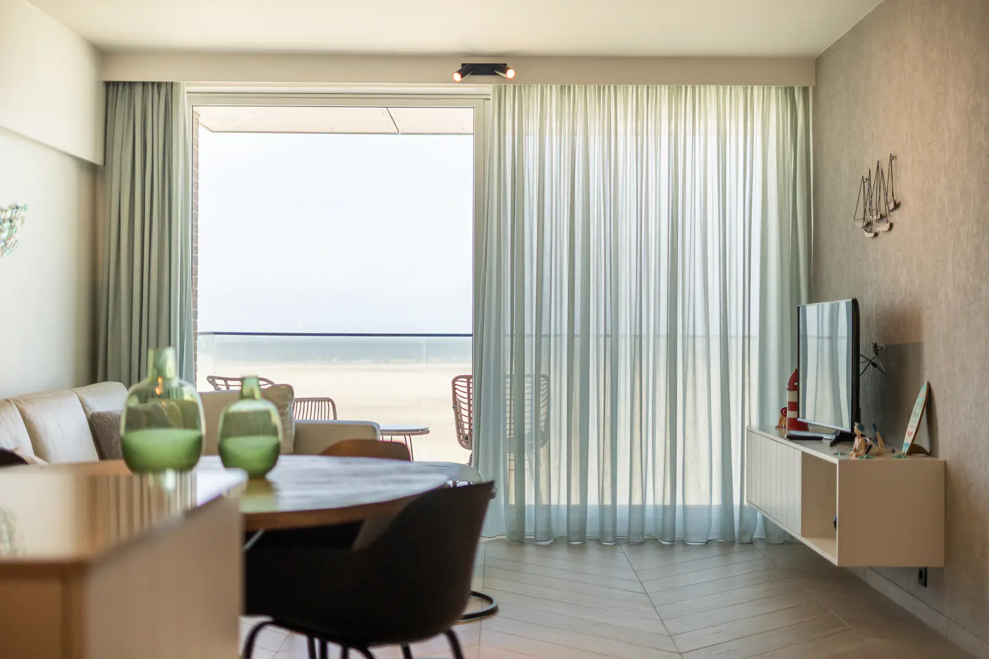 Living room with ocean view. Sheer curtains, wood table, and TV on a white console. Neutral tones.