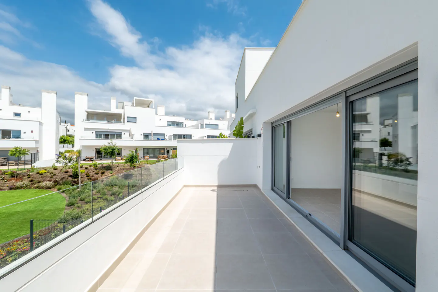 Modern white balcony with glass railings overlooking a landscaped garden and white residential buildings under a blue sky.