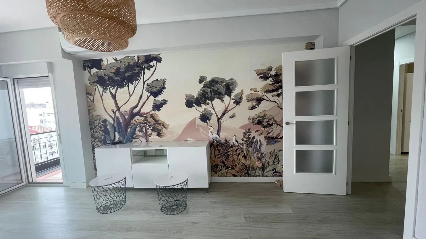 Modern room with a nature-themed mural, white sideboard, two wire stools, a woven pendant light, and a frosted glass door.