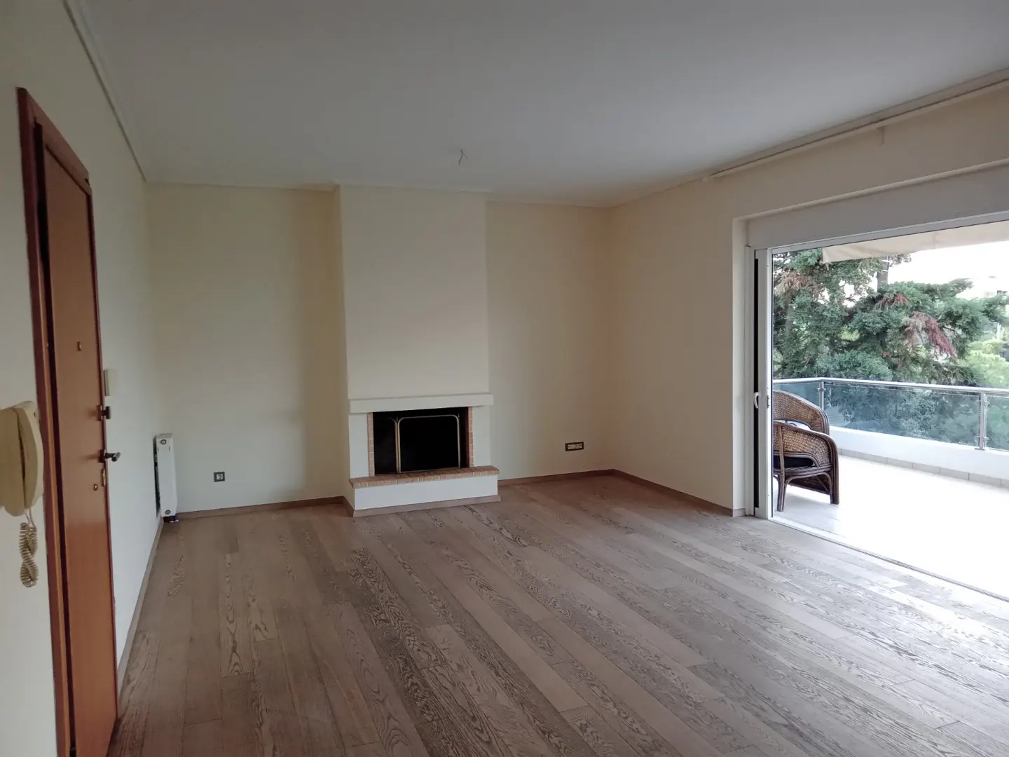 Empty living room with wood floors, fireplace, and balcony with a chair. Walls are off-white.